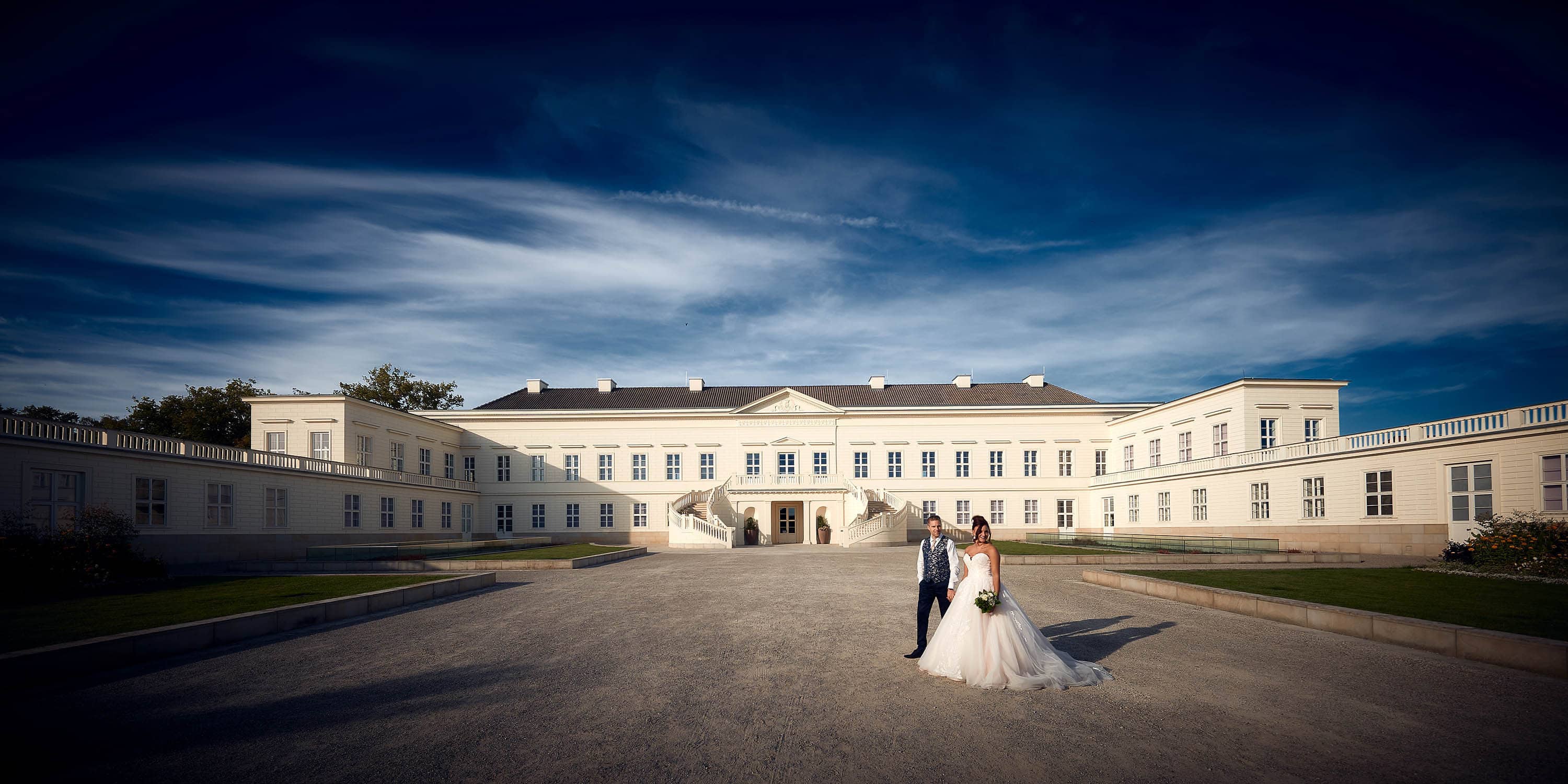 Brautpaar steht bei blauem Himmel vor dem Schloss - Herrenhäuser Gärten Hannover © Hochzeitsfotograf www.hochzeitsverliebt.de